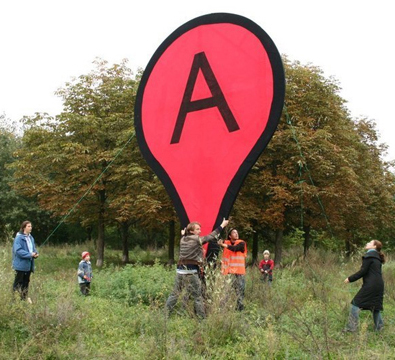 A giant pin shaped sign with the letter 'A' and loads of people holding it up.