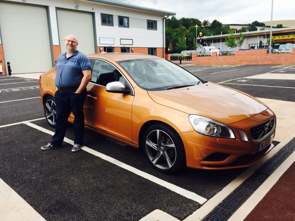 A man smiling, stood next to a bronze Volvo car in a car parking bay.