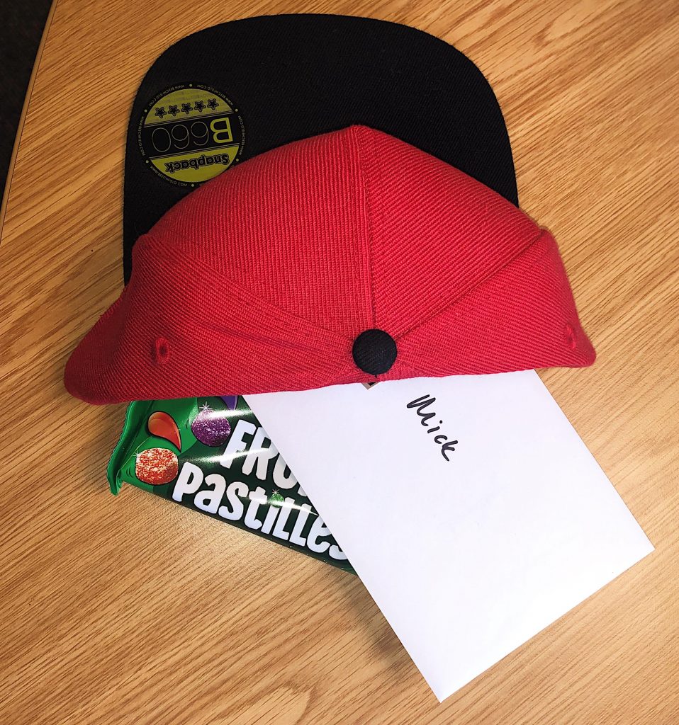 A red and black baseball cap, packet of fruit pastilles and a white envelope saying 'Mick'.